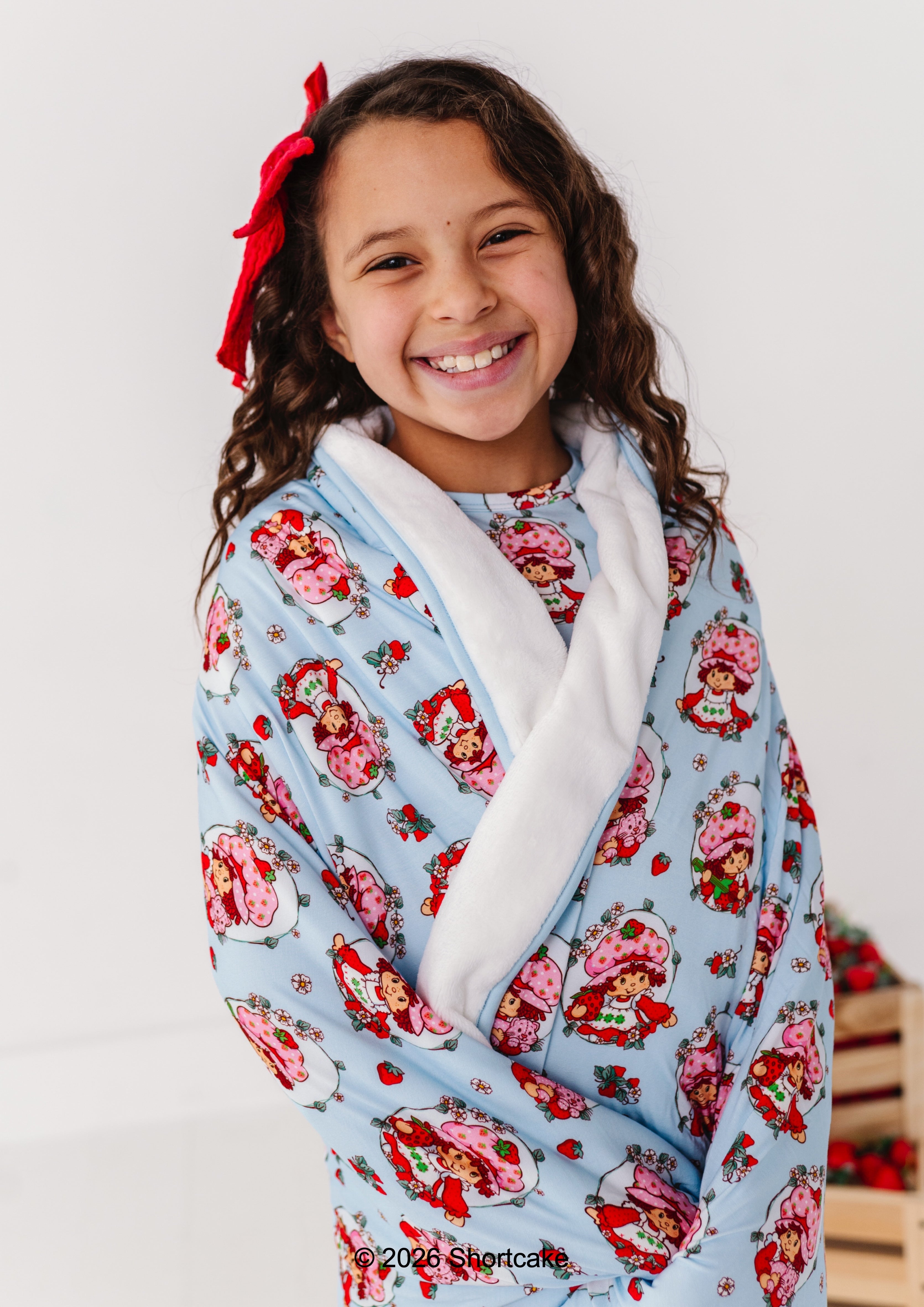Child wrapped in a Strawberry Shortcake™ plush bamboo blanket, smiling against a plain background